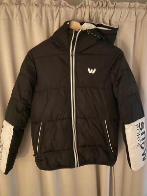 Svart pufferjacka Whistler med huva - Svart pufferjacka från Whistler med vit huva och vita detaljer på ärmarna där det står 'SNOW FORCE'. Jackan har dragkedja framtill, två fickor med vita dragkedjor och en stor logga på huvan. Använd en vinter, Inga märkabara defekter alls! storlek 176 men passar nog även större.