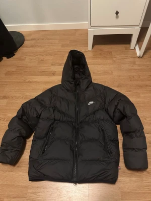 Svart pufferjacka från Nike med huva - Svart pufferjacka från Nike med huva och två stora fickor med dragkedja. Jackan har en clean look med diskret Nike-logga på bröstet och quiltad design. Perfekt för kalla dagar och har en skön, oversized passform.priset kan sänkas 