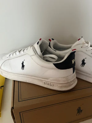Vita sneakers från Polo Ralph Lauren - Snygga vita sneakers från Polo Ralph Lauren med klassisk logga på sidan och mörk blå detalj vid hälen. Skorna har snörning, platt sula och diskreta röda och blå detaljer på hälen och plösen. Det är storlek 42 men passar även runt 41
