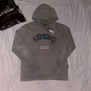 Grå hoodie från Hoodrich - Säljer en grå hoodie från Hoodrich i storlek M. Tröjan har svart och blå broderad text framtill och stort tryck på ryggen. Klassisk huva och känguruficka. Materialet är mjuk bomullsmix, perfekt för streetwear-stil.
