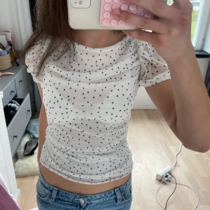 Vit prickig blus med puffärmar - Söt vit blus med svarta prickar och puffiga korta ärmar. Blusen har rynkade detaljer och en figurnära passform. Perfekt för dig som gillar en feminin och trendig stil.