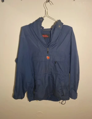 Vintage Fjällräven anorak S - Anoraken är stor i storleken!!! Säljer en blå vindjacka från Fjällräven med huva, halv dragkedja och klassisk logga på bröstet. Jackan har stora fickor framtill, justerbar midja och muddar vid ärmslut. Perfekt för dig som vill ha en stilren och funktionell jacka till vardagen.