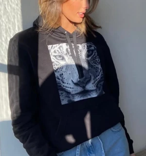 Svart hoodie med leopardtryck - Svart hoodie med stor leopardbild framtill och snörning i huvan. Tröjan har en klassisk känguruficka och är långärmad. Perfekt för dig som gillar djurmönster och vill ha en chill och snygg stil.