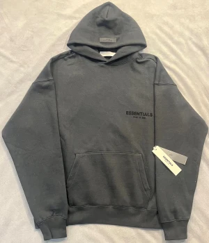 Svart Essentials Fear of God hoodie - Svart hoodie från Essentials Fear of God med stor logotyp på ryggen och mindre tryck på bröstet. Tröjan har huva, känguruficka och ribbade muddar. Materialet är mjuk bomull och passformen är loose, perfekt för en chill streetwear-look.