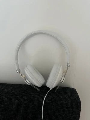 Vita kabelanslutna over-ear hörlurar - Stilrena vita over-ear hörlurar med kabel och 3,5 mm kontakt. Inbyggd mikrofon och fjärrkontroll på sladden. Mjuka öronkuddar för bekväm användning. Hörlurarna är i gott skick utan synligt slitage eller defekter.