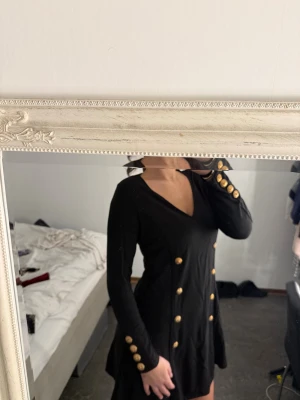 Svart kort klänning med guldknappar - Snygg svart kort klänning med v-ringning och långa ärmar. Klänningen har dekorativa guldknappar längs framsidan och på ärmsluten, vilket ger en lyxig känsla. Passformen är figurnära upptill och kjolen har lite vidd. Perfekt för dig som gillar statement-detaljer.