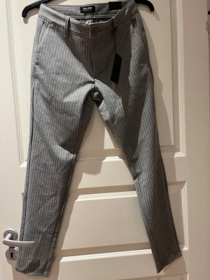 Grå randiga kostymbyxor Only & Sons - Snygga grå kostymbyxor från Only & Sons med smala vita ränder. Byxorna har normal passform, bälteshällor, framfickor och bakfickor med knapp. Perfekta för en clean och stilren look. Materialet är mjukt och bekvämt.