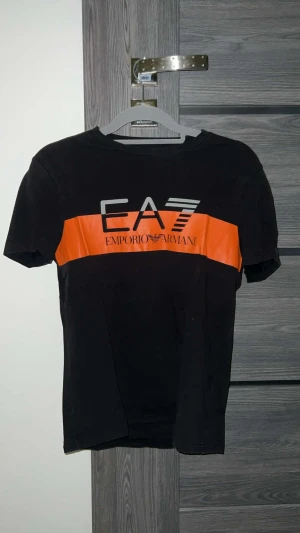 Svart EA7 Emporio Armani t-shirt - Svart t-shirt från EA7 Emporio Armani med en bred orange rand över bröstet och stor logga. Klassisk rund hals och korta ärmar. Materialet är mjuk bomull som känns skönt mot huden. Perfekt för dig som gillar streetwear och snygga märkesplagg.