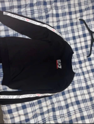 Svart EA7 sweatshirt med logoband - Svart sweatshirt från EA7 Emporio Armani med vita logoband längs ärmarna och tryckt EA7-logga på bröstet i vitt och rött. Tröjan har rund hals och lång ärm, perfekt för en sportig och stilren look. Materialet är mjuk bomull med inslag av polyester.