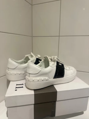 Valentino Rockstud sneakers vit/svart - Snygga Valentino Rockstud sneakers i vitt skinn med svart band och ikoniska pyramidnitar. Använda cirka 5 gånger och mycket bra skick. Säljer då dem tyvärr är för små. Lådan har tyvärr ett hål från frakten dock!Märkeslogga på plösen och innersulan. Perfekta för dig som gillar exklusiv streetstyle.