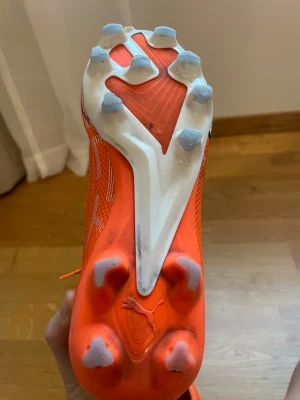 Orange fotbollsskor från Puma - Säljer ett par grymt snygga orangea fotbollsskor från Puma med vita och svarta detaljer. Skorna har snörning och en platt sula med dobbar för extra grepp på planen. Ovandelen är i syntetmaterial med mesh för bra ventilation och lätt känsla.