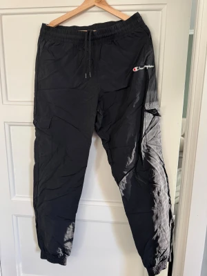 Svarta cargopants från Champion - Svarta cargopants från Champion med broderad logga på benet. Byxorna har elastisk midja med snörning, praktiska fickor med lock på sidorna och resår vid bensluten. Perfekt för en chill och sportig stil. Materialet känns lätt och smidigt. Behöver strykas och textil lim till märket annars är byxorna som nya!