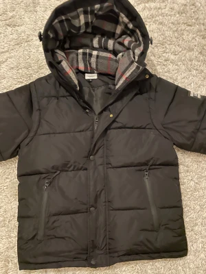 Svart pufferjacka med avtagbara ärmar - Svart pufferjacka från Burberry med avtagbara ärmar, så du kan använda den som väst. Jackan har huva med grått, rött och svart rutigt foder, två fickor med dragkedja och knäppning framtill. Perfekt för kalla dagar och riktigt snygg stil.