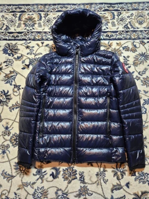 Mörkblå dunjacka Canada Goose - Säljer en mörkblå dunjacka från Canada Goose med glansig finish, huva och två långa dragkedjor framtill. Jackan har det klassiska Canada Goose-märket på ärmen och quiltad design. Perfekt för kalla dagar och har en riktigt snygg look.