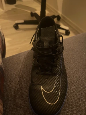 Svarta Nike fotbollsskor med blå sula - Säljer ett par svarta Nike fotbollsskor med blå platt sula, perfekt för inomhusspel. Skorna har snörning, ett stort vitt Nike-märke på sidan och ribbad textur på ovansidan. Materialet är syntet och designen är sportig och modern.