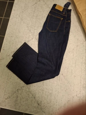 Mörkblå jeans från Nudie Jeans, W30/L34 - Säljer ett par klassiska mörkblå jeans från Nudie Jeans med snygga kontrastsömmar. Modellen har raka ben och fem fickor. Tillverkade i slitstarkt bomullstyg och passar perfekt till en clean och stilren look.
