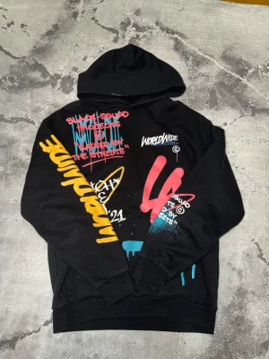 Black Squad graffiti (New Yorker) - Svart hoodie från Black Squad med relaxed fit och huva. Tröjan har färgglada graffiti-prints i gult, blått, rött och vitt både fram och bak, med texter som 'DRIP2HARD' och 'WORLDWIDE'. Tillverkad i mjuk bomull, perfekt för dig som gillar streetwear och bold vibes.