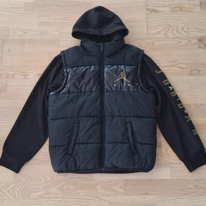 Svart pufferjacka från Jordan Air 12-13 år - Svart pufferjacka från Jordan Air med huva och dragkedja. Jackan har guldigt Air Jordan-logga på bröstet och tryck på ena ärmen. Materialet är polyester och jackan har två sidofickor. Perfekt för dig som gillar sportig streetstyle.