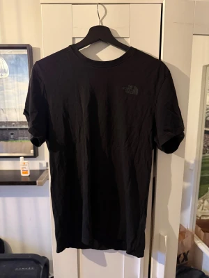 Svart t-shirt från The North Face - Svart t-shirt från The North Face med klassisk rund hals och korta ärmar. Framsidan har en diskret logga, medan baksidan har ett stort tryck i gult och grått med texten 'Never Stop Exploring' och 'The North Face'. Tillverkad i mjuk bomull för skön känsla.