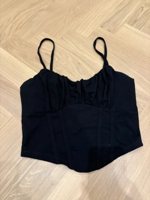 Svart korsett-topp från Dilvin - Svart korsett-topp från Dilvin med smala axelband och markerade sömmar framtill. Toppen har en lätt rynkad överdel och är croppad i modellen. Perfekt för att lyfta vilken outfit som helst.