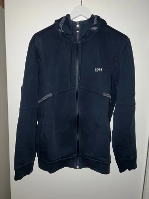 Hugo BOSS hoodie med dragkedja - Snygg mörkblå hoodie från BOSS med vit logga på bröstet. Tröjan har huva med snörning, hel dragkedja framtill och ribbade muddar. Detaljer med mönstrad snörning vid huvan och axlarna ger en cool touch. Perfekt för chill dagar eller när du vill vara extra bekväm. Säljer den på grund av att den är för stor för mig. Har använt den max 2 gånger. Den kostade 1800kr, så priset på 900kr är väldigt långt med tanke på det.