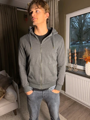Grå hoodie med dragkedja och huva - Säljer en stilren grå hoodie med dragkedja och huva. Tröjan har snörning i huvan, två fickor framtill och ribbade muddar vid ärmslut och nederkant. Perfekt för chill dagar och enkel att matcha med jeans eller joggers.