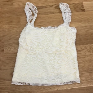 Vit spetslinne med rosett och volang - Supersött vitt linne i spets med volangkant. Toppen har ett blommigt spetsmönster och en liten rosett framtill vid halsringningen. 