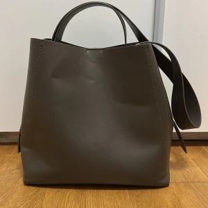 Brun axelväska från Gina Tricot, bra skick. - Snygg brun axelväska från Gina Tricot i stilren och minimalistisk design. Väskan är tillverkad i syntetmaterial med slät yta och har både handtag och axelrem. Insidan har en praktisk ficka med dragkedja. Perfekt storlek för att få plats med allt du behöver.