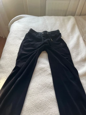 Svarta mjukisbyxor Juicy Couture - Svarta mjukisbyxor från Juicy Couture med broderad JC-logga på fickan och snörning i midjan. Byxorna är i mjukt fleecematerial och har en klassisk loose passform som är riktigt skön. Perfekta för chill dagar eller när du vill ha en avslappnad look.