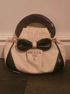 Vit handväska från Prada med rosett - Snygg vit handväska från Prada med svart rosettdetalj i croco-imitation. Väskan har silverfärgade metalldetaljer, dragkedja och klassisk Prada-logga framtill. Insidan är fodrad med mönstrat tyg med Prada-tryck. Perfekt för dig som gillar lyxiga accessoarer. Väskan har söndrig dragkedja och därav priset💕 men funkar enkelt att stänga med hjälp av knappen🎀 finns inget serienummer som jag kan hitta💕