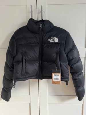 Svart croppad pufferjacka North Face - Säljer en svart croppad pufferjacka från The North Face med broderad logga på bröstet. Jackan har hög krage, dragkedja framtill och två sidofickor med dragkedja. Puffig design och justerbar nederkant för extra stilpoäng.