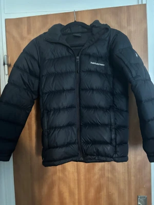 Svart Peak Performance dunjacka - Säljer en svart dunjacka från Peak Performance med huva och diskret logga på bröstet och ärmen. Jackan har quiltad design, dragkedja framtill och är långärmad. Perfekt för kalla dagar och har en clean, sportig look. Har bara andvänts i 1 vinter