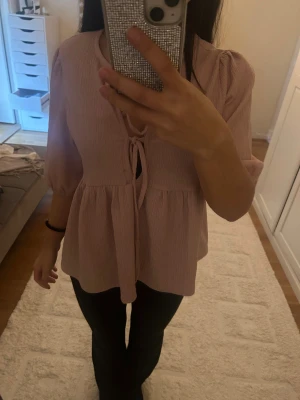 Ljusrosa blus med knyt och puffärm - Supersöt ljusrosa blus med knyt framtill och puffiga halvlånga ärmar. Blusen har en lätt rynkad, flowy passform och är tillverkad i ett tunt, strukturerat tyg. Perfekt för dig som gillar romantiska och feminina detaljer.nästan aldrig använd 