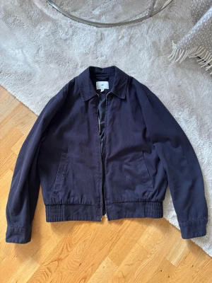 Mörkblå jacka från H&M - Snygg mörkblå jacka från H&M med klassisk krage och ribbade muddar vid midjan. Jackan har dragkedja framtill och två sidofickor. Materialet är bomull och jackan är långärmad med knappdetalj vid ärmslutet. Perfekt för dig som gillar stilren och enkel design.