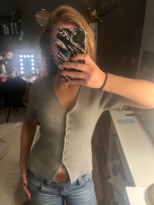 Grå stickad topp från Gina  - Säljer en grå stickad topp med v-ringning och korta ärmar. Toppen har en rad snygga knappar framtill och en liten slits nedtill som ger en cool detalj. Passformen är slim och materialet känns mjukt och bekvämt mot huden. Perfekt att styla med jeans eller kjol.