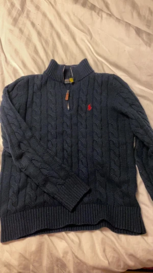 Marinblå stickad tröja Polo Ralph Lauren - Säljer en marinblå stickad tröja från Polo Ralph Lauren med klassiskt kabelstickat mönster och röd logga på bröstet. Tröjan har half zip i halsen med läderdetalj och ribbade muddar vid ärmslut och nederkant. Perfekt för dig som gillar stilren och tidlös design.