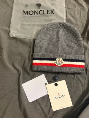 Grå Moncler mössa med ränder - Snygg grå ribbstickad mössa från Moncler med klassiska ränder i marinblått, vitt och rött på uppviket. Mössan har Moncler-logga framtill och är tillverkad i mjukt ullmaterial. Perfekt för dig som vill ha en clean och exklusiv look.