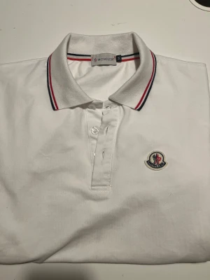 Vit pikétröja från Moncler med logga - Vit pikétröja från Moncler med klassisk krage och knäppning framtill. Kragen har snygga ränder i rött och marinblått, och tröjan pryds av Monclers broderade logga på bröstet. Tillverkad i mjuk bomull som är skön mot huden. Ny pris: 650kr