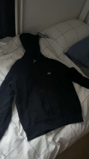 Lacoste hoodie svart - Svart hoodie från Lacoste med klassisk krokodillogga på bröstet. Tröjan har huva med snörning och dragkedja framtill. Perfekt för dig som gillar en clean och stilren look. Långa ärmar och mjukt material gör den extra skön.