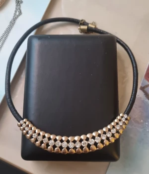 Svart halsband med guld och strass - Snyggt halsband med svart band och dekorativt parti i guldton med glittrande strass. Designen är modern med runda guldfärgade detaljer och gnistrande stenar som ger extra bling. Perfekt för att lyfta din outfit med lite extra glamour.