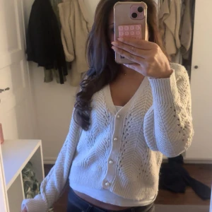 Vit stickad kofta med pärlknappar - Superfin vit stickad kofta med v-ringning och pärlknappar framtill. Koftan har ett snyggt hålmönster och ribbade muddar vid ärmslut och nederkant. Perfekt att slänga över ett linne för en chill look. Köpt på sellpy för 249kr