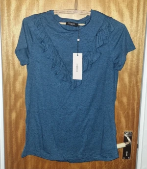 Blå topp med volang från Soaked - Säljer en snygg blå topp från Soaked med korta ärmar och volangdetalj i V-form framtill. T-shirten har en mjuk och skön känsla, perfekt för dig som gillar stilrena plagg med en twist. Passar dig som vill ha något enkelt men ändå lite extra.