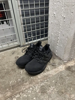 Svarta Adidas Ultraboost sneakers - Säljer ett par helsvarta Adidas Ultraboost sneakers med stickad ovandel och klassiska tre ränder på sidan. Skorna har snörning, platt sula och rund tå. Boost-sulan ger grym komfort och passar perfekt för dig som gillar stilrena och sportiga sneakers.