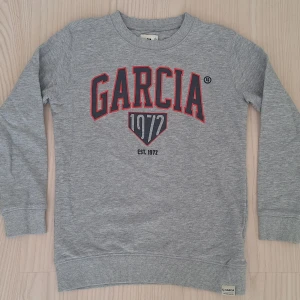 Ny Grå sweatshirt från Garcia stl 152-158 - Snygg grå sweatshirt från Garcia med tryck i marinblått och rött framtill. Tröjan har rund hals, långa ärmar och ribbade muddar vid ärmslut och nederkant. Perfekt för en avslappnad och sportig look.
