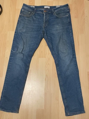 Slim Glenn blå jeans storlek 34/32 - Snygga blå jeans från Jack & Jones i modellen Slim Glenn. Jeansen har en slim passform, klassisk femficksdesign och en liten sliten detalj på vänster ben. Tillverkade i mjukt denimtyg med en mellanblå tvätt och subtila slitningar.