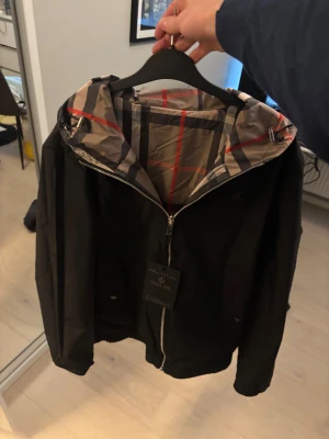 Svart vindjacka från Burburry med huva - Svart vindjacka från Burburry med dragkedja och huva. Insidan av huvan har ett rutigt mönster i beige, svart och rött. Jackan har långa ärmar, två sidofickor och en stilren look som passar perfekt till streetwear.