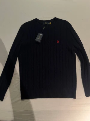 Polo Ralph Lauren tröja - Snygg mörkblå stickad tröja från Polo Ralph Lauren i storlek XXL. Tröjan har kabelstickat mönster, rund hals och lång ärm. Klassisk röd Polo-logga broderad på bröstet. Perfekt för dig som gillar stilrena och tidlösa plagg.