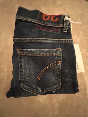 Dondup blå jeans med slitningar - Snygga mörkblå jeans från Dondup med coola slitningar och färgstänk på både fram- och baksida. Klassisk femficksmodell med raka ben och låg midja. Jeansen har detaljerade sömmar och en tagg i midjan. Perfekt för dig som gillar en edgy streetstil.