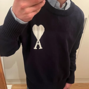 Mörkblå stickad tröja från Ami - Snygg mörkblå stickad tröja från Ami med ikoniskt vitt hjärta och A på bröstet. Rund hals, ribbade muddar och klassisk passform. Perfekt att styla med skjorta under för en clean look. Materialet är mjukt och håller värmen under kyliga dagar. Jag köpte den begagnat här på Plick och då var den helt ny så skicket är väldigt bra då jag alldrig använt den. Om ni har några frågor så svarar jag så snabbt jag kan. 