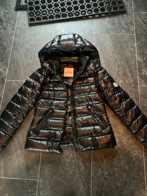Svart ÄKTA Moncler pufferjacka - Säljer en svart ÄKTA pufferjacka från Moncler med glansig finish och huva. 💛Jackan har två snedställda fickor med dragkedja, Moncler-logga på vänstra ärmen och quiltad design. Har tyvärr inget kvitto men QR koden på bild 4 visar att den är äkta. 🥰Perfekt för dig som vill ha en snygg och varm jacka med exklusiv känsla.💕